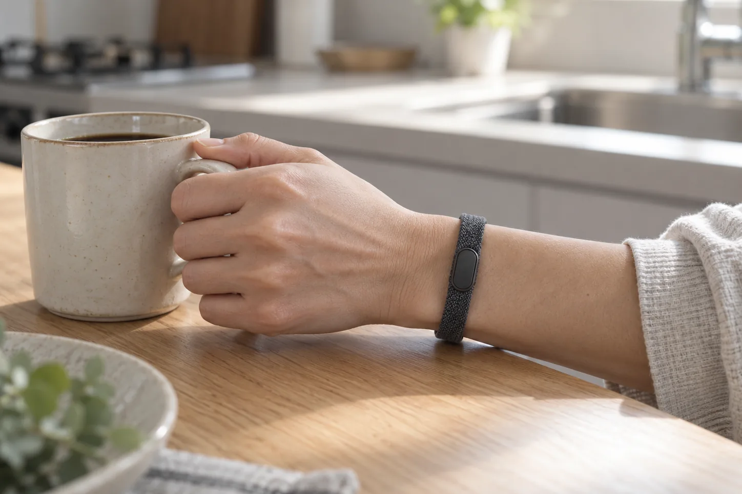 キッチンでマグを持つ手。手首に、画面のない細い Fitbit のバンド。